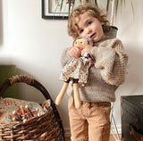 A young child stands beside a basket, wearing a handknit merino wool sweater in a neutral colour called Freshwater Pearl. He is hugging a teddy bear plushy in a dress